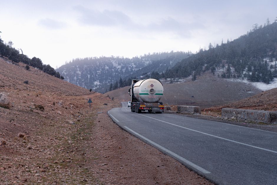 Gas tanker truck traveling on a mountainous road with snow-capped hills in the background.