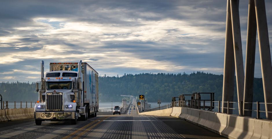 A semi-truck travels across a bridge in Washington, USA, with cloudy skies and forested landscape.