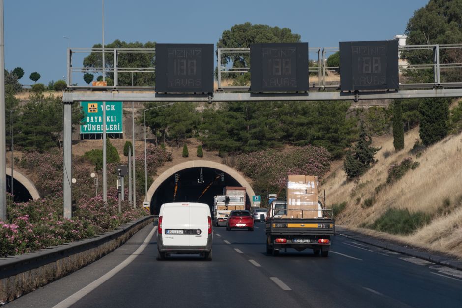 View of vehicles on a highway approaching the Bayraklı Tunnel in İzmir, Türkiye, with road signs and greenery.